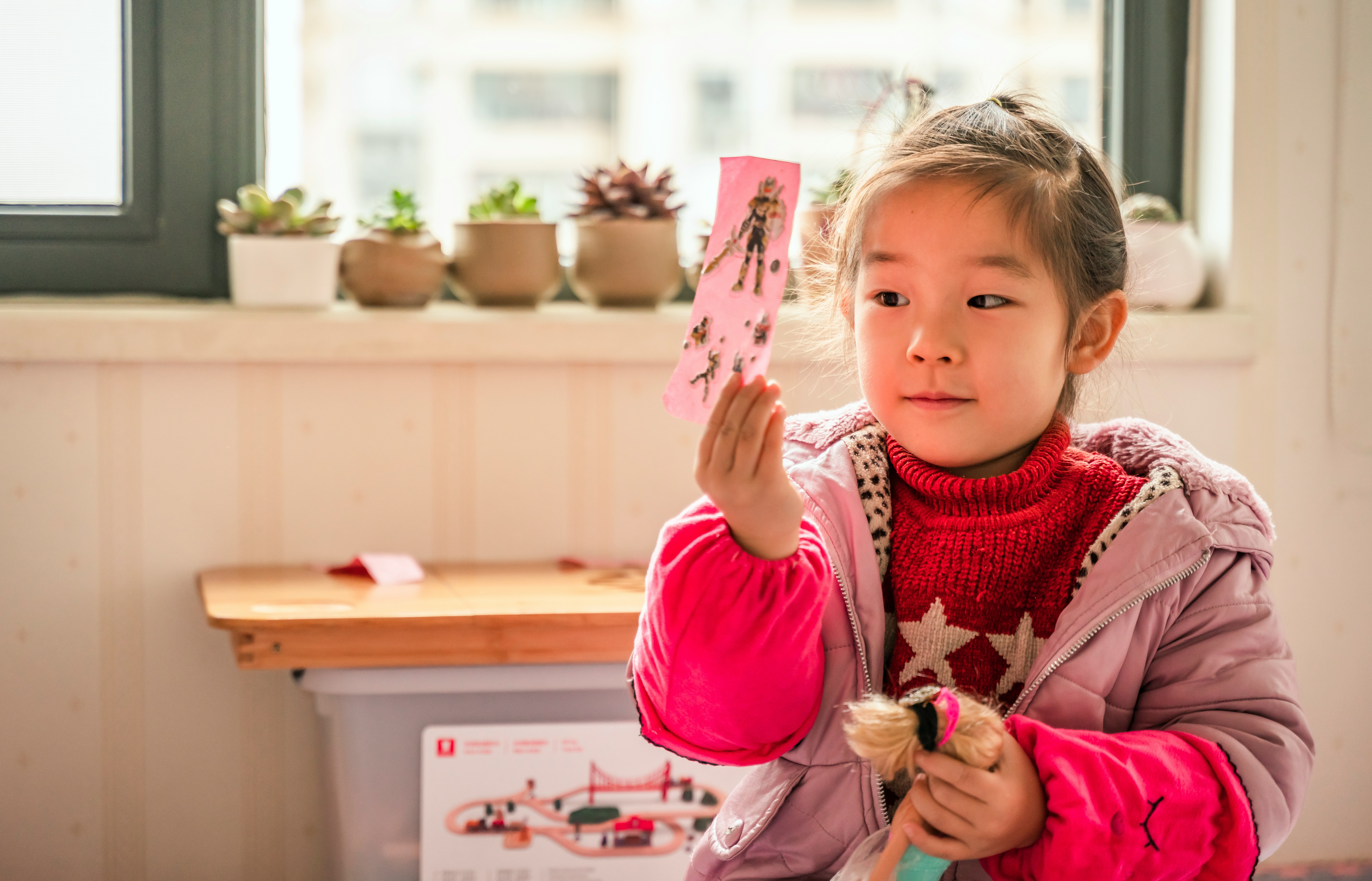 A little girl holds a pink card with stick figure people on it.