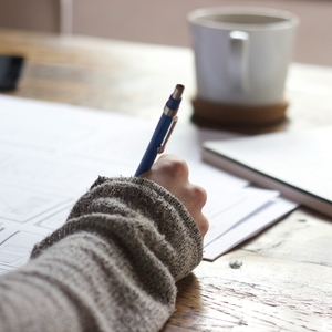 Person writing with pen on a desk with notebook and mug.